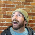A blurry image of radio host, Jonny J with a big grin looking up at the ceiling. He's wearing a beanie and is standing in front of a red brick wall